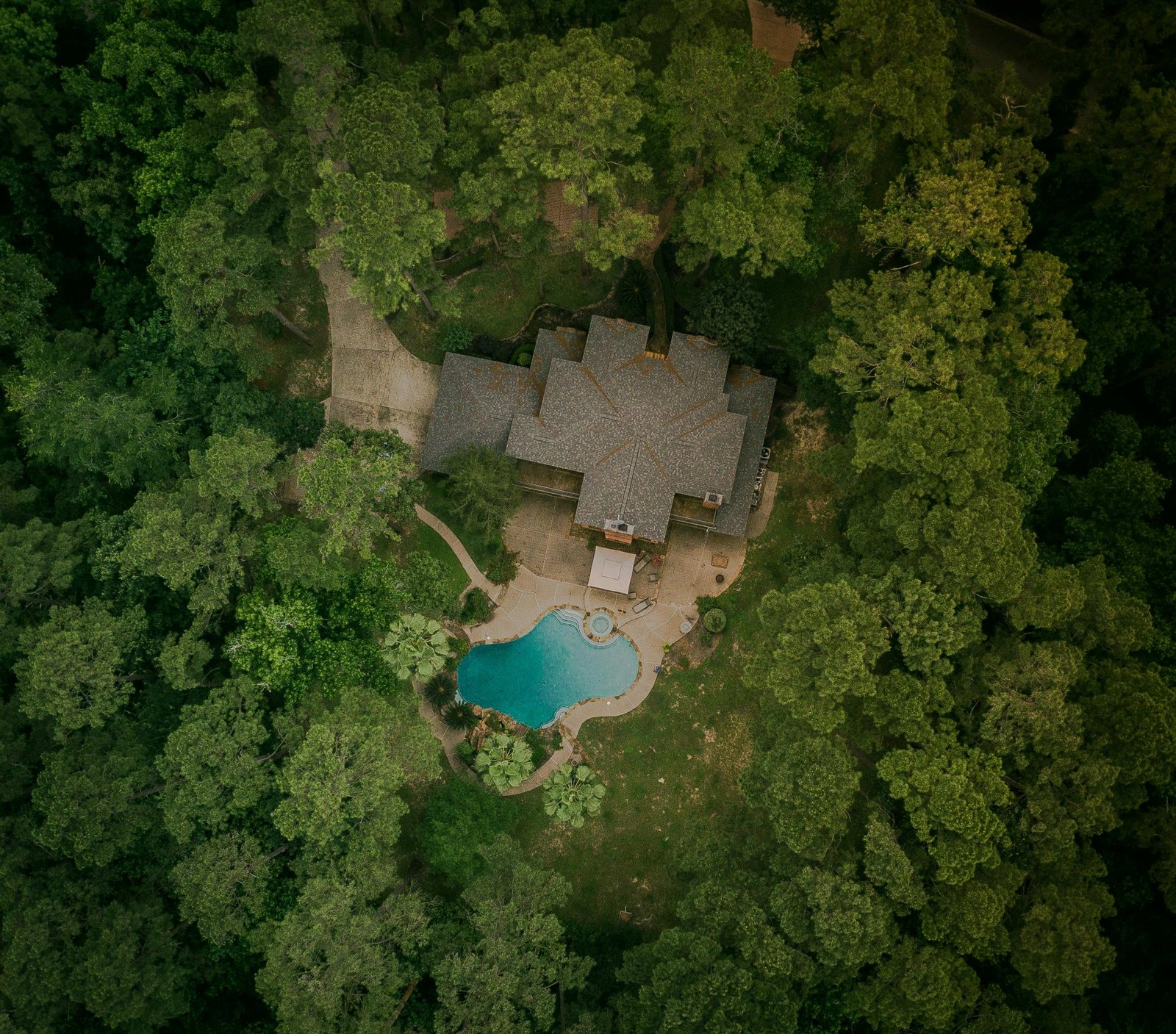 A scenic aerial shot of a house surrounded by lush forest and a swimming pool.